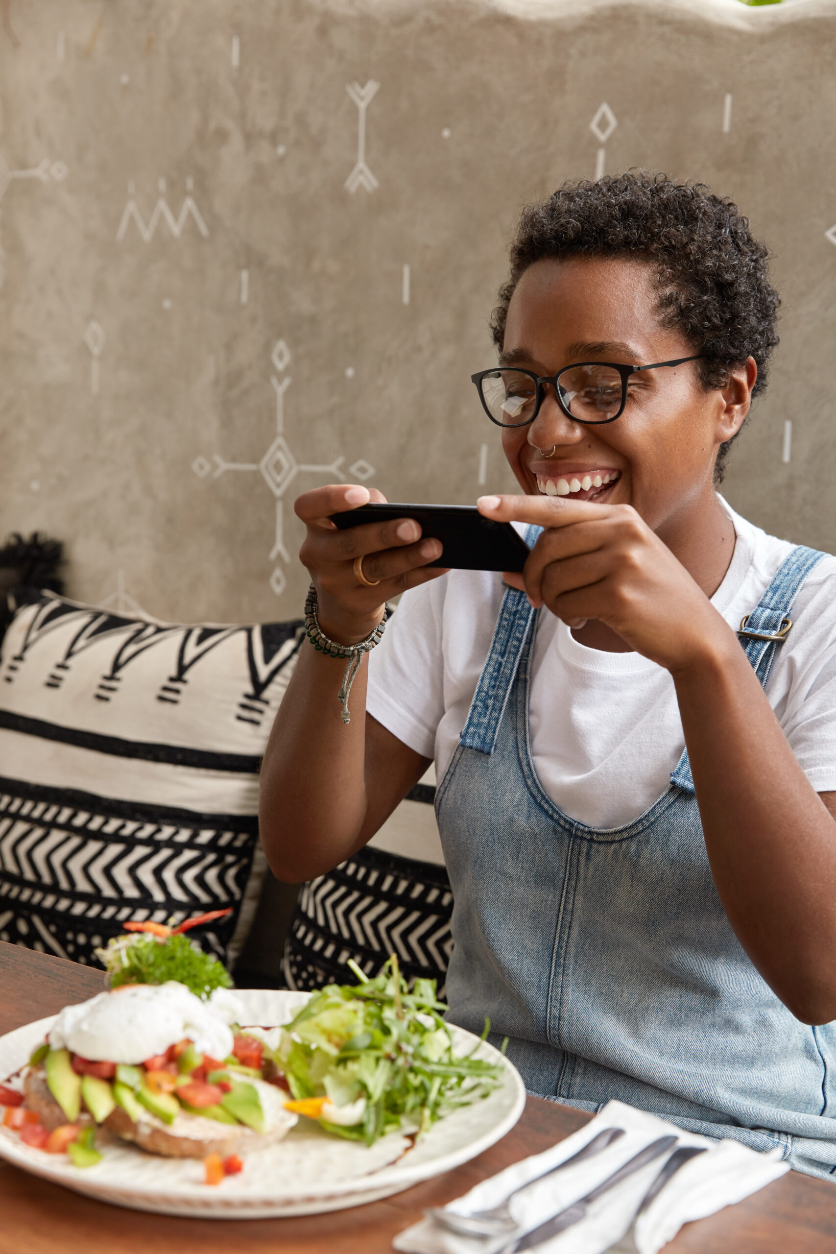 Young woman smiling while using a food delivery app on her smartphone, seated at a restaurant table with a healthy meal in front of her.