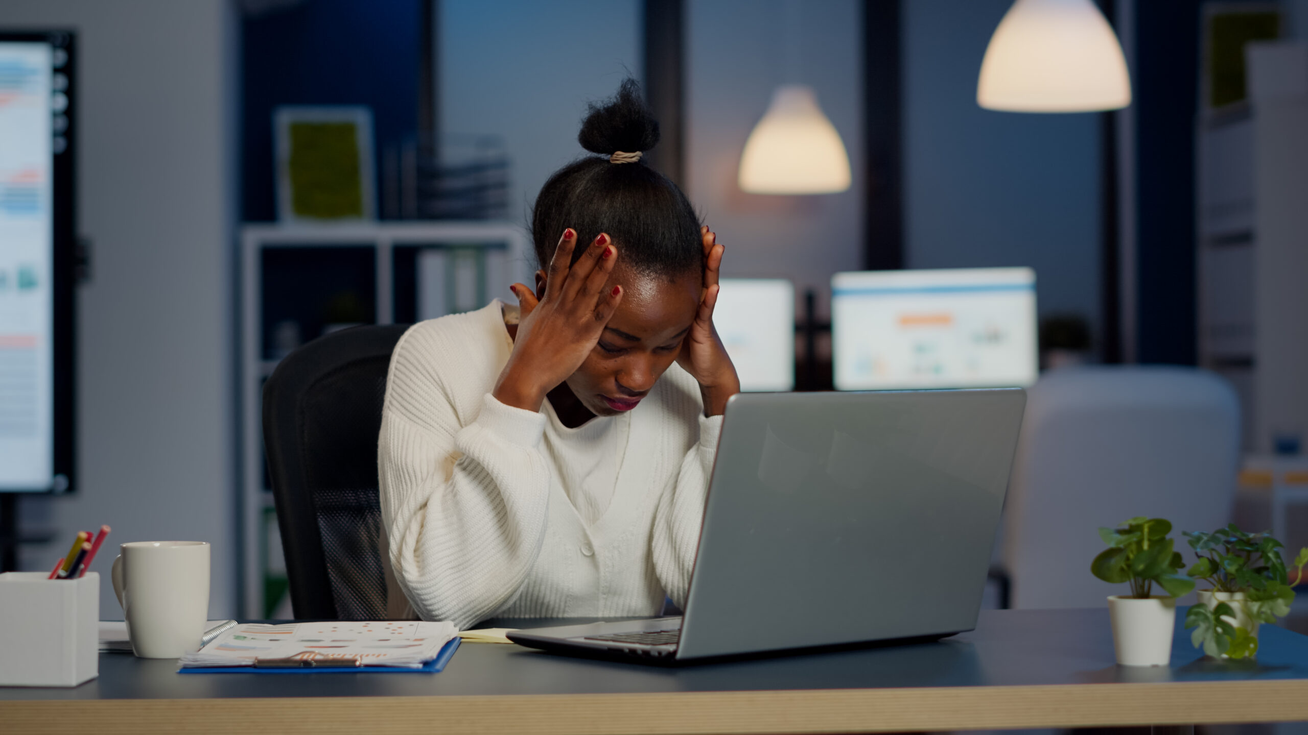 A stressed Black businesswoman holding her head in her hands while working late at night in a dimly lit office, with a laptop, documents, and a coffee mug on her desk.