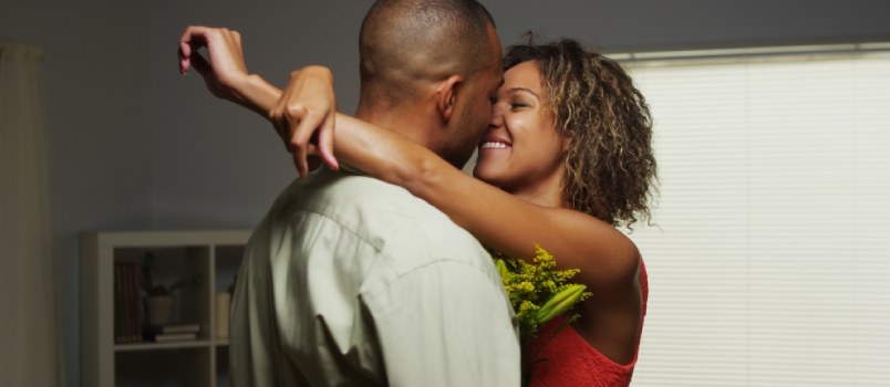 "Couple embracing closely indoors, woman in red dress holding flowers, smiling romantically"