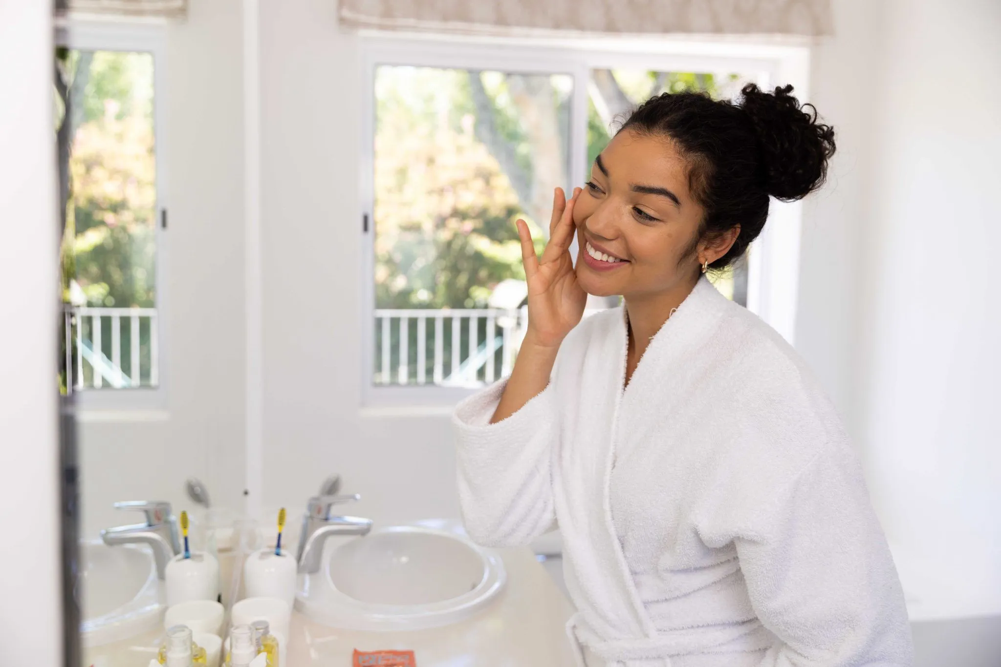 Woman in a white bathrobe applying skincare in front of a bathroom mirror during her morning routine in a bright, modern home.