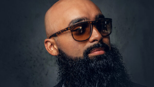 Close-up portrait of a bald man with a full beard wearing dark sunglasses in a studio setting.