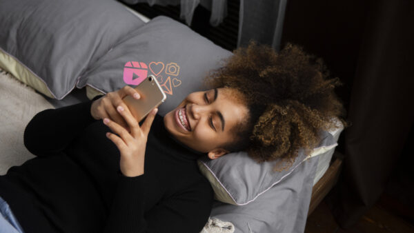 Young woman smiling while lying on a bed and using her smartphone, with social media icons floating above the screen to represent online engagement and messaging.