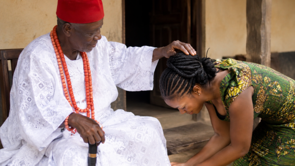 Young Nigerian woman respectfully bowing before an elderly man in traditional clothing.