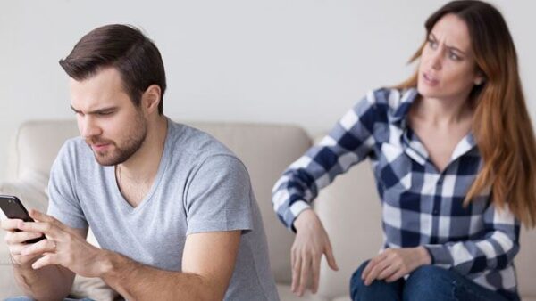 A woman looks frustrated as a man sits beside her and focuses on his phone.