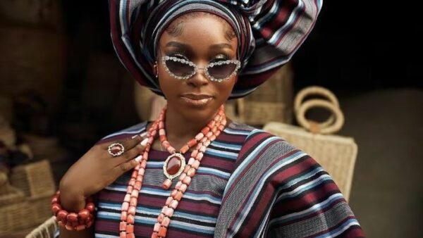 Nigerian woman wearing a striped Aso Oke outfit and gele with coral beads and modern accessories.